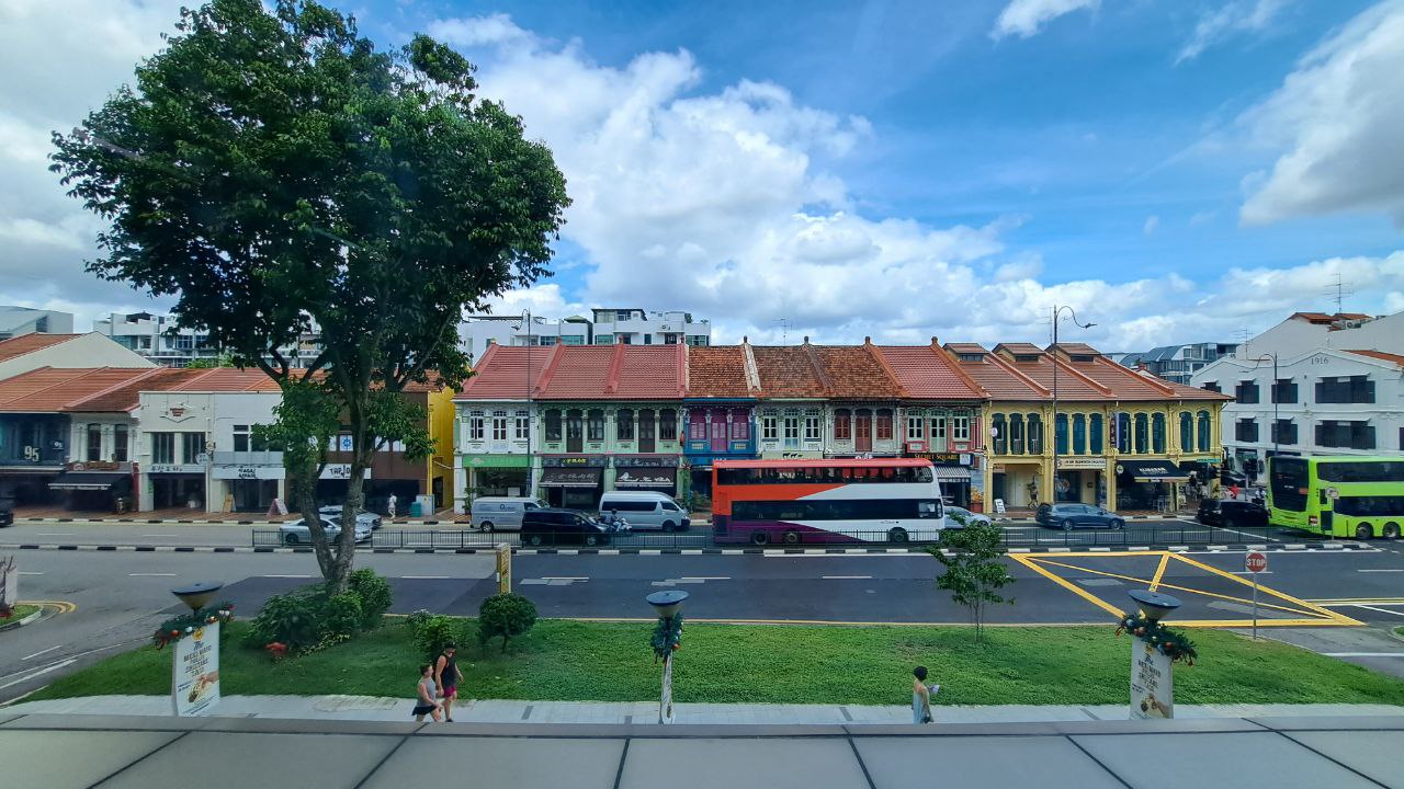 a red and white doubledecker bus is stopped in the middle of a street in an urban setting. there are colorful shophouses and buildings on either side of the street. the sidewalks are lined with trees and there are people walking in the distance. the day is sunny and the sky is blue.
