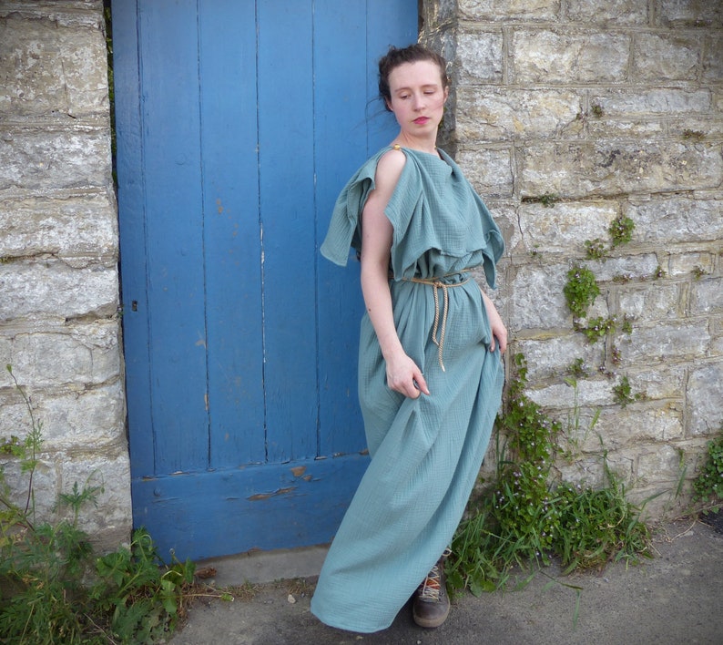 a woman is standing outside next to a blue door and a stone wall. she is wearing a flowing teal dress that appears to be inspired by ancient greek or roman styles.  the dress has a loose, comfortable fit and is cinched at the waist with a rope belt.  she is wearing brown boots. plants grow at the base of the stone wall. the overall setting suggests a historical or periodinspired photoshoot.
