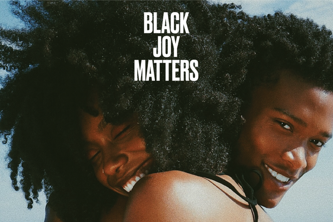 the image showcases two young black women, likely friends or sisters, embracing and laughing together on a sunny beach. the woman in the front has her arm around the other's shoulder, while the woman in the back leans her head on the other's, both with joyful smiles. their natural hair is styled in large afros. the text "black joy matters" is overlaid in white capital letters, emphasizing the importance of black happiness and wellbeing. the overall atmosphere is vibrant and positive, celebrating black joy.