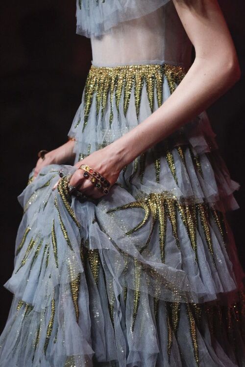 a woman in a beautiful blue tulle gown with gold sequins and a sparkling bodice. she is holding the dress in her hands.  she is wearing several rings with colorful stones.