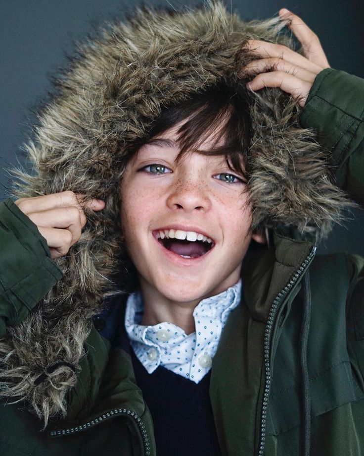 a young boy with brown hair wears a green hooded winter coat with a fur lining and white shirt with blue polka dots. he is smiling and laughing with his hand on the fur hood of his coat.