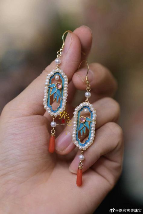 a woman's hand is holding a pair of beautiful, vintage, chinese earrings. the earrings feature an ornate design with turquoise enamel, pearls, and coral. the woman's hand is in focus, showcasing the intricate details of the jewelry. the background is blurred, highlighting the earrings.
