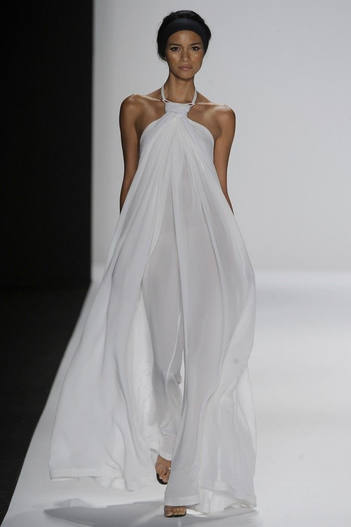a model is walking on a runway wearing a long, white dress. the dress is simple and elegant, and it flows beautifully as she walks. the model has black hair and she is wearing high heels. she is posing for the cameras. the runway is white, and the background is a white wall.