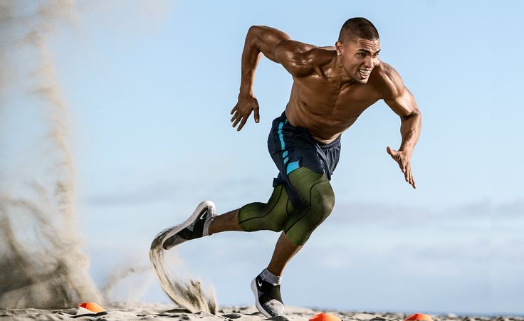 a muscular man in athletic attire is sprinting on a sandy beach. his face is focused and determined as he pushes himself to run faster. the sand kicks up behind him creating a dust cloud.  he is in excellent shape, clearly dedicated to his fitness. there are orange cones in the background, suggesting a training routine.
