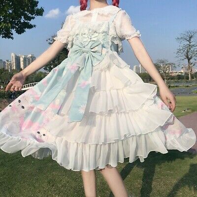a woman wearing a flowing blue and white lolita dress. the dress is tiered and has a floral pattern and lace accents. the woman is also wearing a white lace blouse underneath the dress. she is standing outside on a sunny day. the background of the image shows a grassy area with trees and buildings in the distance.