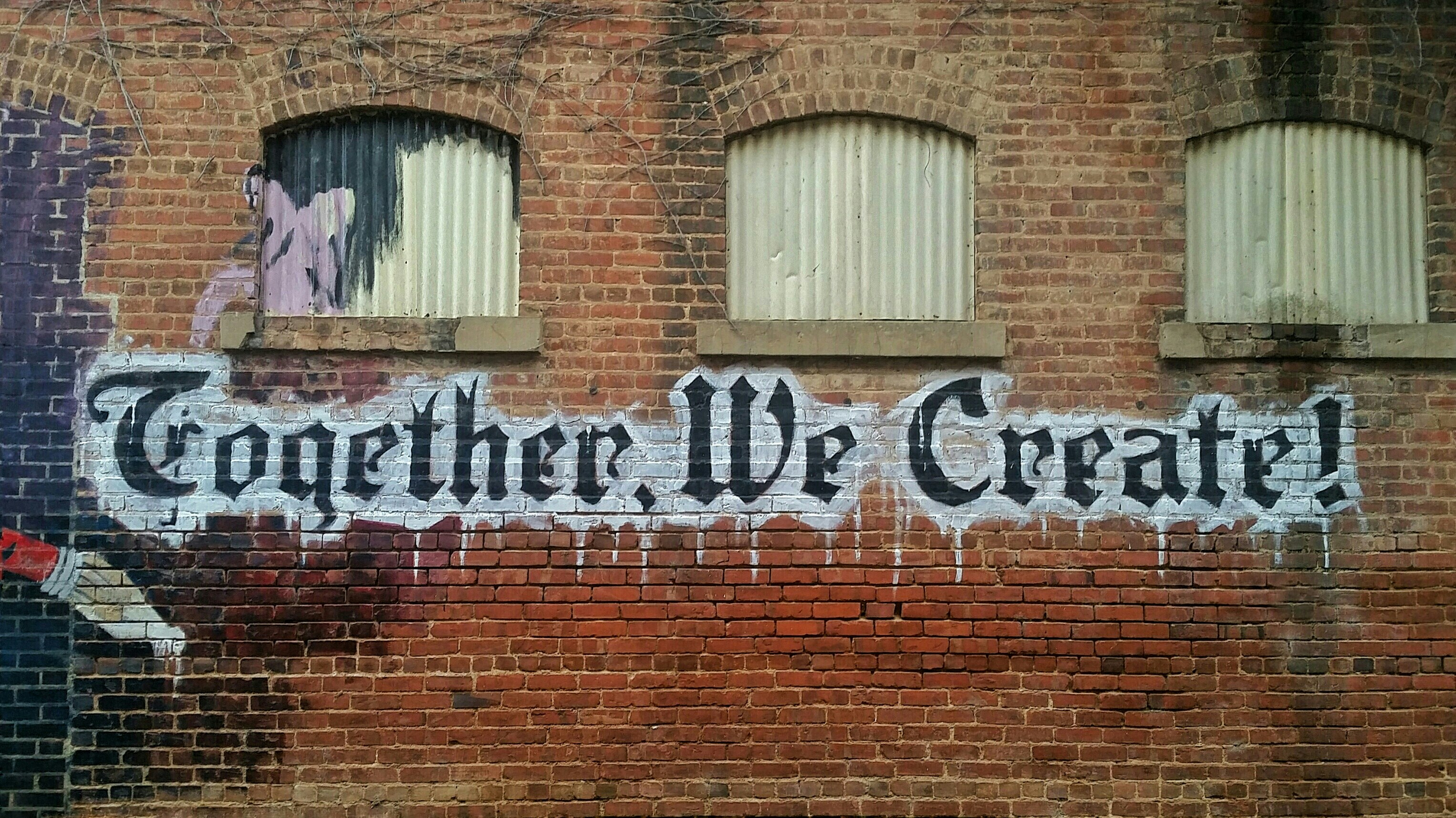 a brick wall with three boardedup windows features the spraypainted words "together, we create" in large, stylized black and white letters.  the wall is a mix of red and brown bricks, and traces of additional artwork are visible on the left side of the image.  the overall tone suggests an urban environment, possibly an older building.