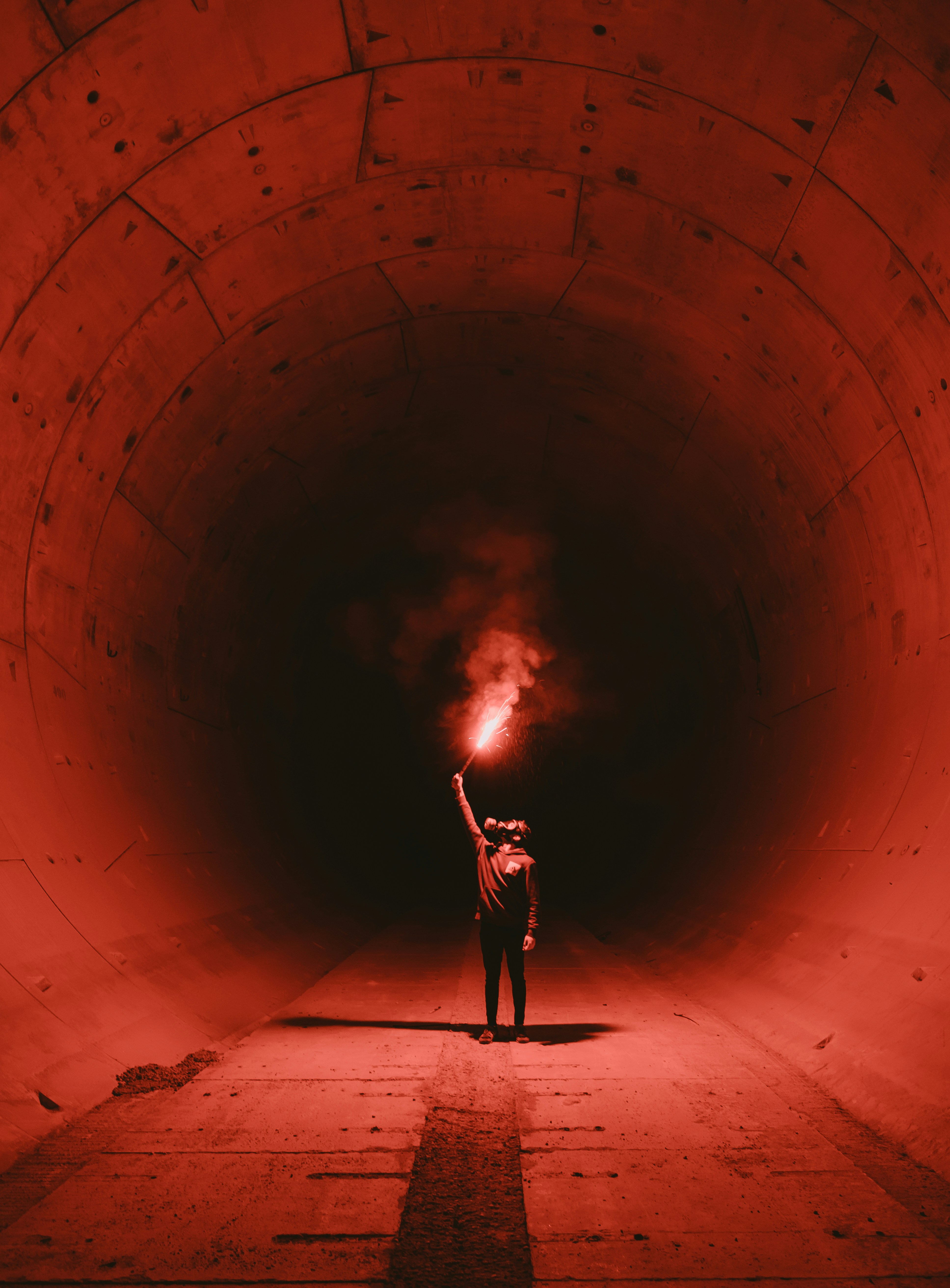 a man stands in the middle of a concrete tunnel illuminated by a red flare he is holding. the tunnel is circular and the perspective is from the ground looking up at the man. the overall tone of the image is dark and mysterious.
