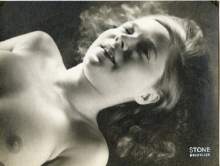 the image is a vintage black and white photograph of a nude woman reclining.  she has long, dark hair and her face is visible, showing a serene or possibly slightly melancholic expression. the lighting is soft and focused on her face and upper body, creating a high contrast against the darker background. the photo appears to be from the early to mid 20th century, potentially the 1920s or 1930s, based on the style.  a small watermark or photographer's mark ("stone bruxelles") is visible in the bottom right corner.  the overall mood is artistic and sensual, emphasizing the beauty of the female form.