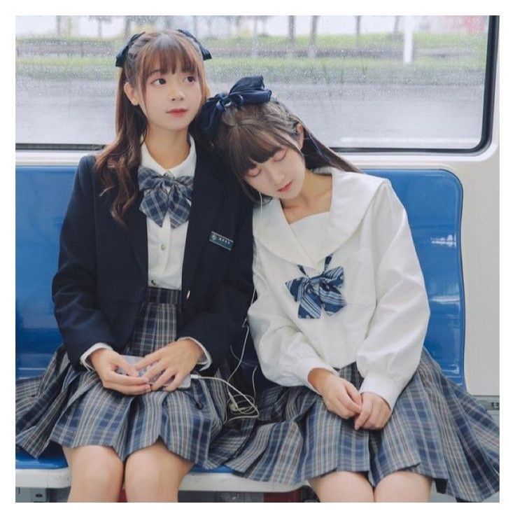 wo young schoolgirls are sitting close together on a train. one girl is wearing a black blazer and a pleated skirt. the other is wearing a white sailor collar shirt and a pleated skirt. both girls are wearing school uniforms and have their hair neatly styled with bows. they appear to be friends and are leaning on each other affectionately. the overall mood of the image is cute and sweet.