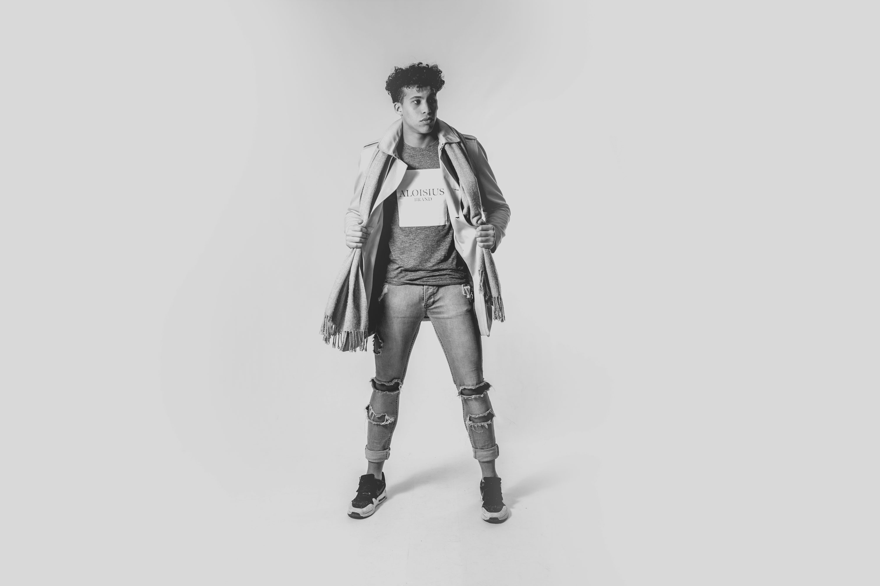 a black and white fulllength studio shot of a young male model standing against a light grey background. he is wearing a grey textured coat draped casually over his shoulders, a grey tshirt with the aloisius brand logo, ripped jeans, and darkcolored sneakers. his pose is relaxed yet stylish, showcasing the clothing. the overall mood is contemporary and fashionable.