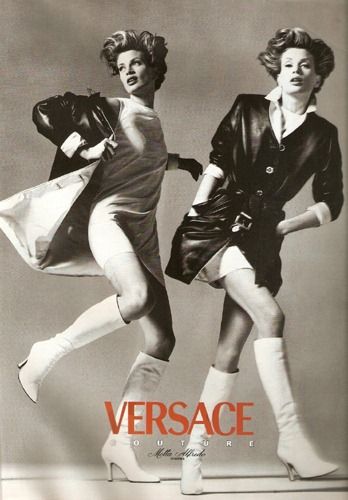 two female models are dressed in fashionable attire. both of the models are wearing leather jackets and white boots. the model on the left has her jacket unbuttoned and is wearing a white dress, while the model on the right has her jacket buttoned and is wearing a black mini skirt. the model on the left is wearing a black fedora hat, while the model on the right is wearing a white top. the models are posing in front of a neutral background, and the image has the versace logo.