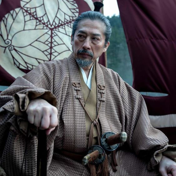 an older man with a serious expression sits in the bow of a boat. he is dressed in traditional japanese clothing and has two swords at his side. the man appears to be a samurai warrior. the image has a muted color palette and soft lighting, suggesting an overcast day.