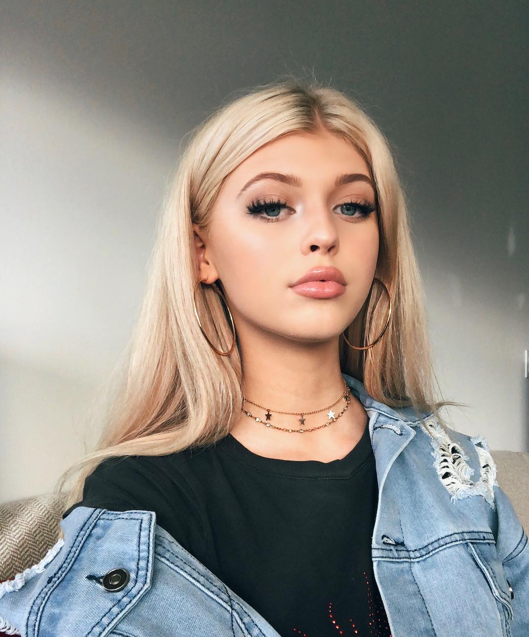 a portrait of a woman with blonde hair, wearing hoop earrings, a necklace, and a denim jacket over a tshirt. she is wearing makeup. it's likely loren gray based on her distinct appearance.