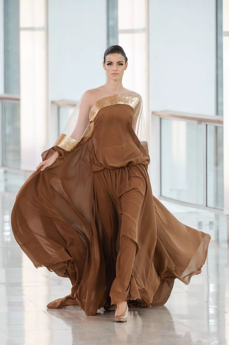 a model walks down a runway in a beautiful brown and gold dress. the dress is flowing and elegant, and the model is posed in a graceful way. the dress is a work of art, and the model is showcasing it perfectly. the dress is a  classic and timeless piece of clothing, and it is clear that it was designed with care and attention to detail.