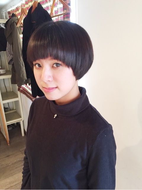a woman with a black bob haircut and a black turtleneck shirt is looking at the camera. she has a serious expression on her face and is standing in front of a white wall.