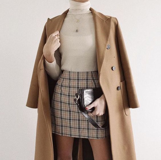 a closeup image showcases a woman wearing a stylish autumnal outfit. the ensemble features a camelcolored long coat draped over a creamcolored turtleneck sweater paired with a brown and beige plaid mini skirt. a small black crossbody bag is held in the woman's hands. the model's overall appearance is sophisticated and preppy, reflecting a minimalist style. the photograph's neutral color palette, combined with the classic elements of the outfit, projects an image of understated elegance.