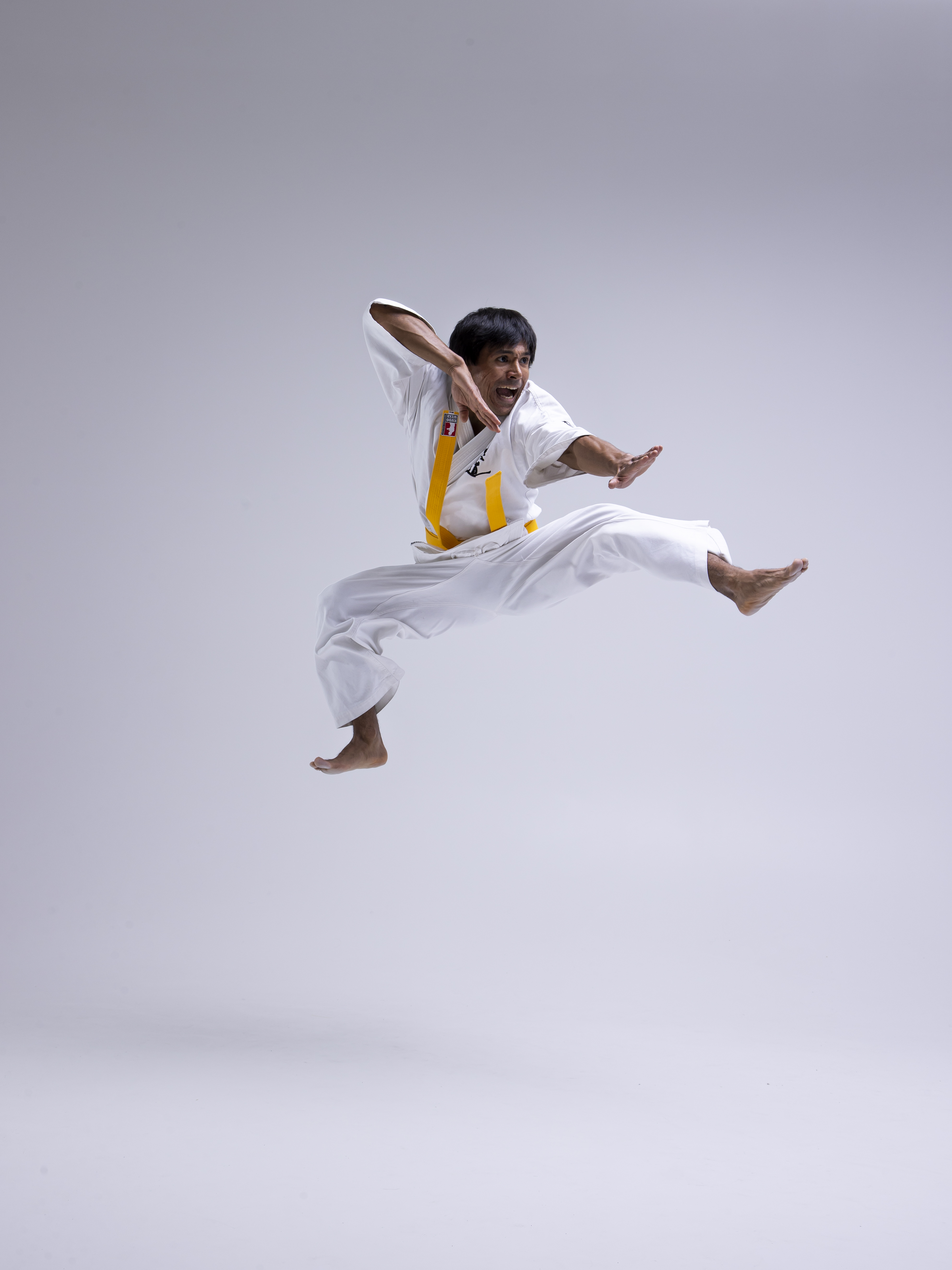 a man in a white karate uniform with a yellow belt is jumping and performing a kick. he is barefoot and appears to be in midair, executing a dynamic martial arts move. the background is a plain light gray.