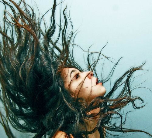 the image captures a woman with long, dark hair in the midst of a dramatic hair flip. her hair is suspended in midair, fanning out around her head against a light blue background. the woman's face is turned upwards, her mouth slightly open as if caught in a moment of exhilaration or release.  the lighting is soft and diffused, creating a sense of movement and fluidity.