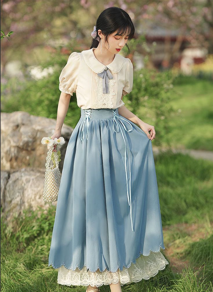 a young woman poses in a garden setting, wearing a vintageinspired outfit. she is wearing a creamcolored blouse with a peter pan collar, puff sleeves, and lace detailing. the blouse is tucked into a highwaisted, flowy blue skirt with lace trim at the hem. the skirt has a tie detail at the waist. the woman is holding a small white purse and has a dreamy expression on her face. the lighting is natural and soft, giving the image a romantic and ethereal feel.