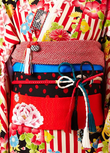 a closeup shot showcases the intricate details of a kimono, focusing on the obi (belt) and the surrounding fabric. the obi is layered with various colors and textures, including a red crosshatched pattern at the top, a solid blue band, a red dotted pattern on a black background, and a solid red band at the bottom featuring a large floral design. a decorative tied knot with multiple colored cords (red, black, teal, and white) hangs from the obi. the kimono itself features a vibrant mix of floral and striped patterns in red, white, black, and other colors.