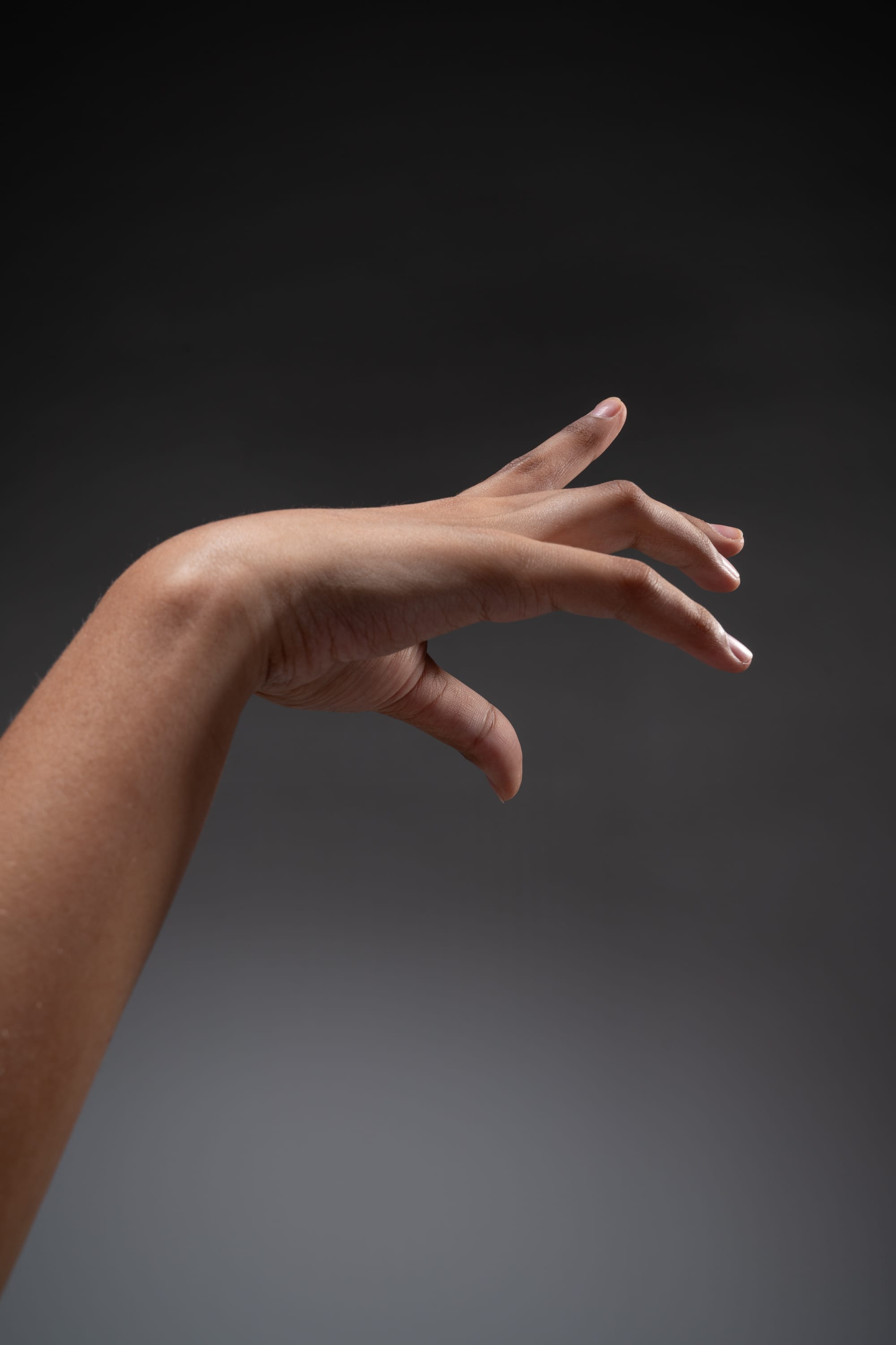 a woman's arm and hand is extended forward against a gray studio background. the hand is open with the fingers slightly curled, as if reaching out or gesturing. the image focuses on the hand and arm, with the rest of the body out of frame. the lighting is soft and even, highlighting the shape and texture of the hand.