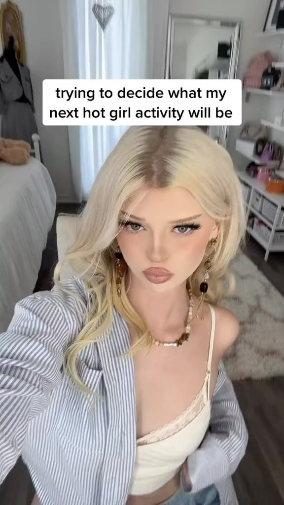 a blonde woman with blonde hair is taking a selfie. she is wearing a white bralette and a striped blue and white shirt. she is also wearing jewelry, including a necklace and earrings. the background is a bedroom. there is text overlaying the image that says "trying to decide what my next hot girl activity will be".