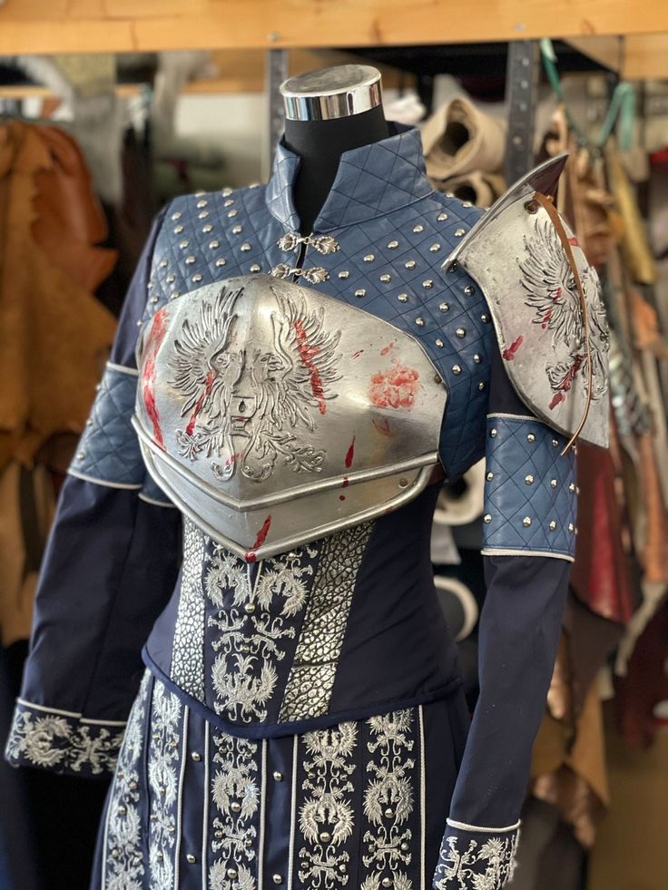 a mannequin displays a blue and silver costume with medieval and fantasy elements. the costume includes a quilted blue bodice with silver studs, a silver chest plate with an eagle design and fake blood spatter, a matching shoulder piece, and a blue skirt and sleeves with silver detailing.