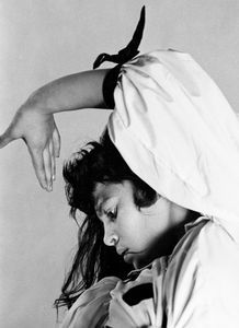 a black and white portrait of a young woman. she is wearing a white dress and a black scarf around her neck. her hair is long and dark. she is looking down and has a thoughtful expression on her face. she is posed in a studio with a simple background.