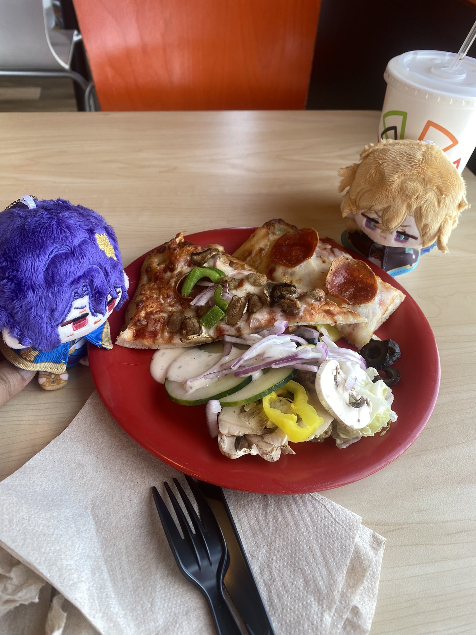 a closeup of a plate of pizza and salad on a wooden table in a restaurant setting. the plate is red and the salad is a mix of green lettuce, red onion, cucumbers, and mushrooms. the pizza is pepperoni and cheese. there is also a fork and napkin on the table. there are plush toys in the background.