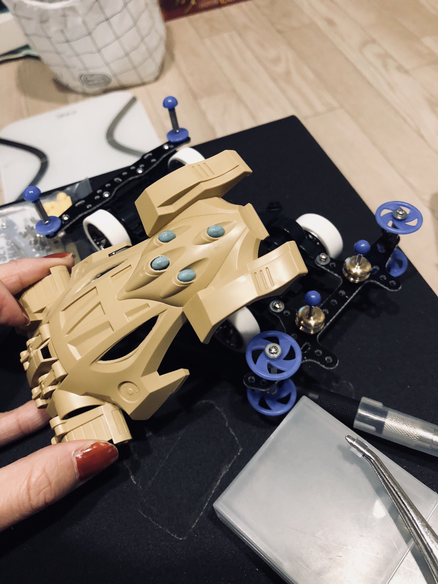 a pair of hands is holding a beige and black toy car body over its chassis and parts. they appear to be in the middle of assembling the toy car. there is a small black container and a set of small tools visible in the image.