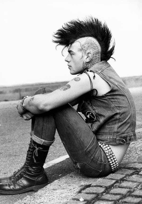 a black and white photo of a punk sitting on the pavement. he has a large mohawk and a dragon tattoo on the side of his head. he is wearing a denim vest, jeans, combat boots, and a studded belt. he is looking off to the side.