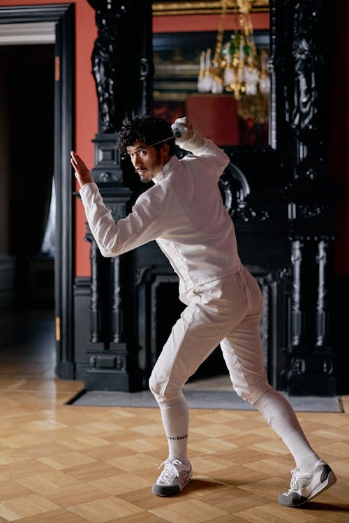 a male fencer, dressed in a white fencing uniform, is captured midaction in a grand, historical interior. he's poised dynamically, his body suggesting a swift movement in a fencing match. the backdrop features a rich, dark woodpaneled room with a classical fireplace. the wood flooring adds to the elegant and historic feel. the fencer's attire is neat and wellfitted, reflecting a focus on both functionality and style. the overall scene blends the dynamism of the sport with the stillness of the grand setting.