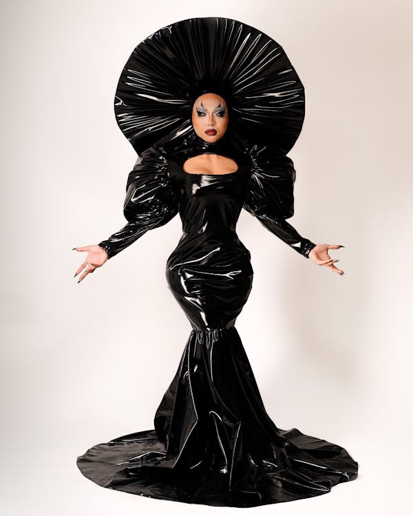 a drag queen stands in a studio wearing a black latex mermaidstyle dress with large puff sleeves and a matching large black hat. the dress features a cutout at the chest. the drag queen has stage makeup on and is extending their arms outwards.