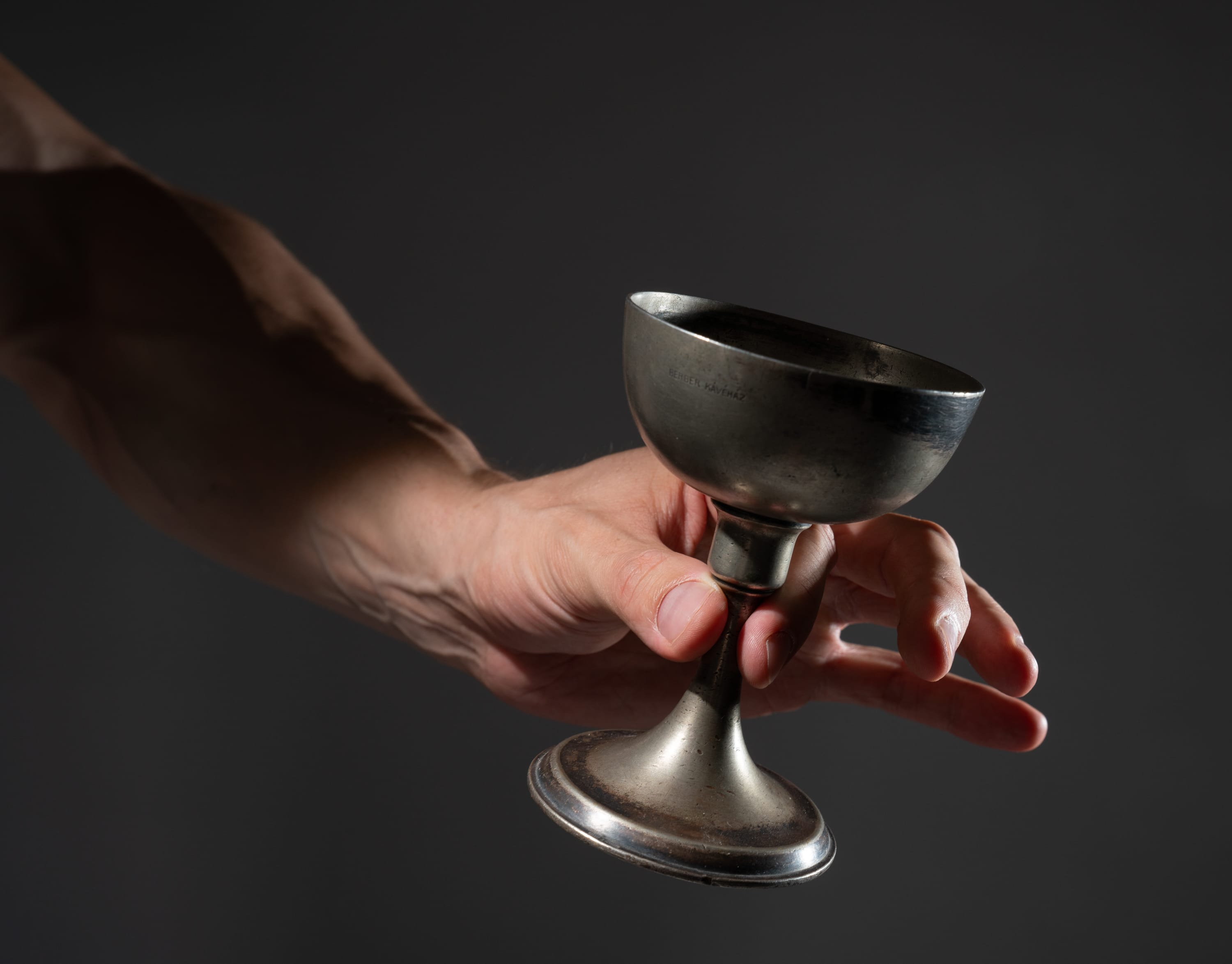 a closeup side view of a fairskinned man's arm and hand carefully holding an antique silver goblet against a dark gray background. the goblet appears to be made of tarnished metal and has a classic chalice shape with a wide bowl and a stemmed base. the man's hand is gently wrapped around the stem of the goblet. the lighting is soft and highlights the texture and details of the goblet and hand.