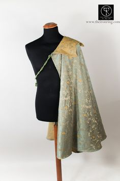 a pale green and gold brocade cloak or cape with a structured shoulder piece, draped over a dress form.  the garment is photographed against a white background in a studio setting.