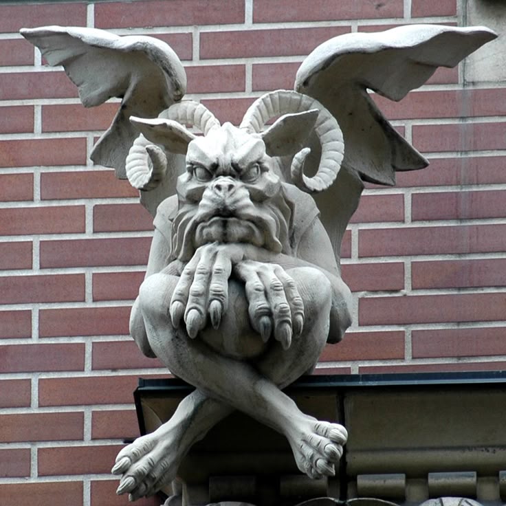 a detailed stone gargoyle sits perched on a building exterior with a brick wall background. the gargoyle is a mythical creature with horns, wings, and a brooding expression. its claws are resting on its crossed legs. the architecture suggests a gothic or medieval style.