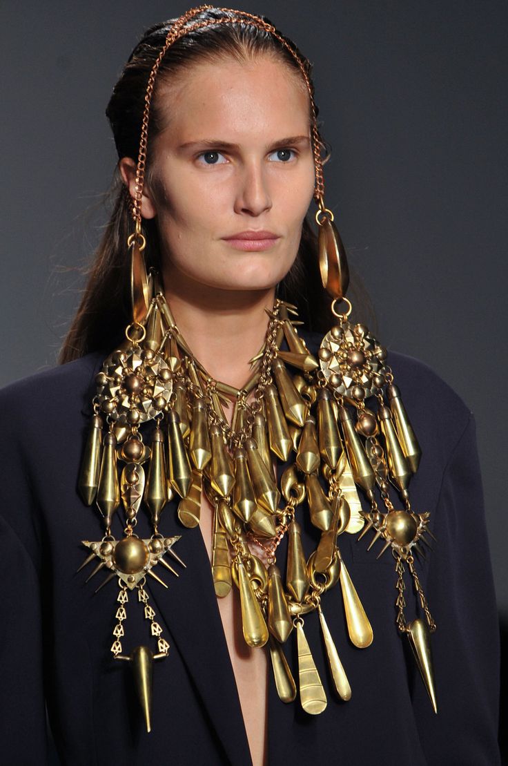 a woman with long dark hair is wearing a gold headpiece, a large statement gold necklace, and gold earrings. she is standing in a fashion show. the necklace is made up of chains, pendants, and other gold embellishments. the earrings are long and dangle, with a large gold disk at the bottom. she is looking down. the background is blurred, but the focus is on the woman and her jewelry.
