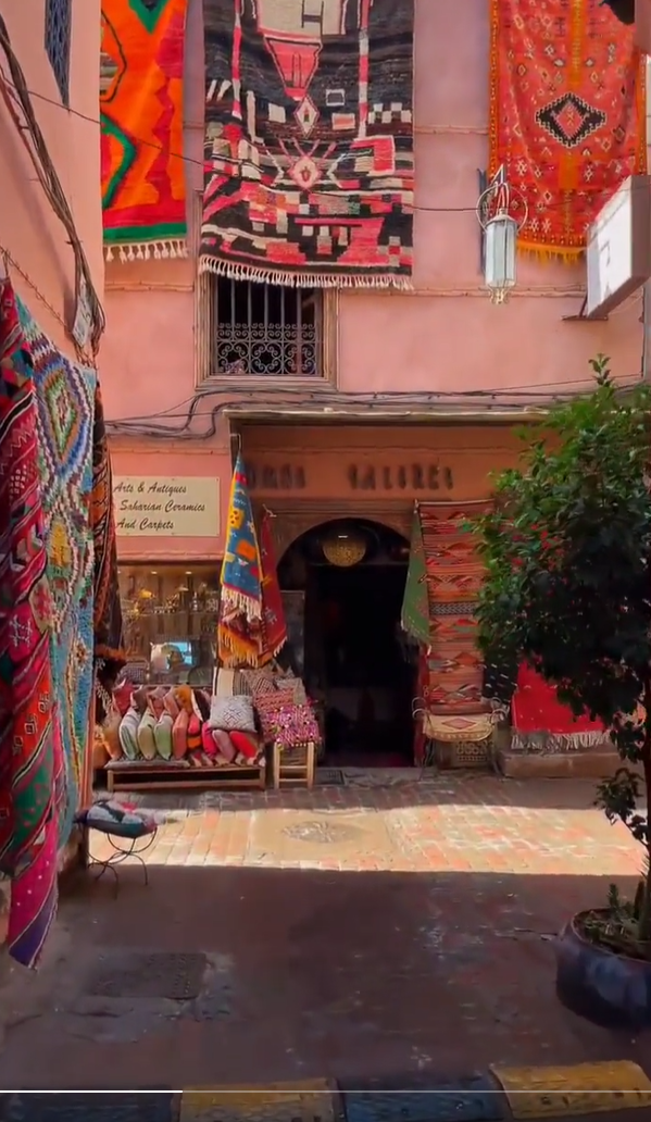 this image shows the exterior of a moroccan store specializing in arts, antiques, ceramics, and carpets. the store is adorned with colorful rugs and textiles hanging on the walls and displayed near the entrance. a variety of items are on display, including carpets, cushions, and handbags. the building has a pink facade with an arched doorway and a sign above indicating it is a gallery. the street is cobblestone and there are some plants in pots nearby.