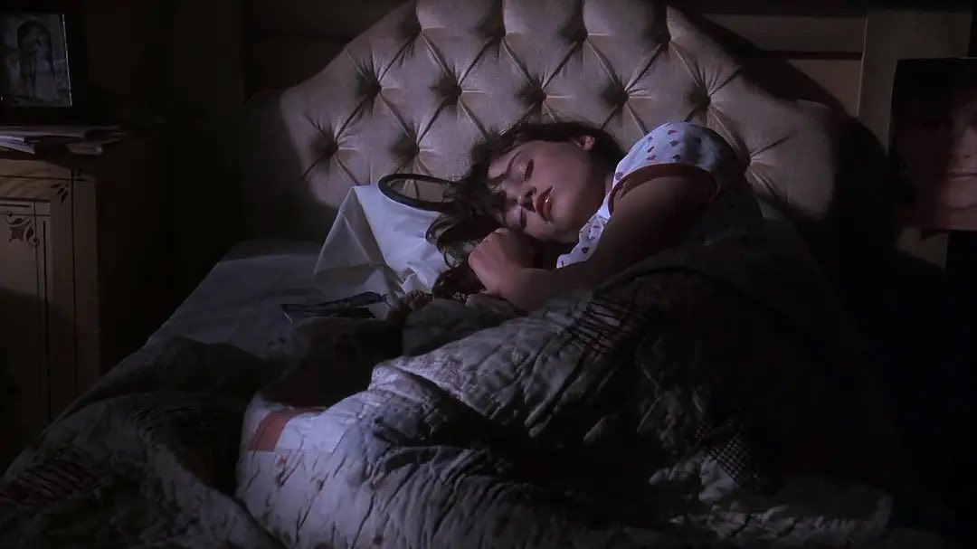 a young girl is asleep in her bed. the room is dark, lit only by a faint light source. she is lying on her side, curled up under a quilt. her dark hair is spread across the pillow. the bed has a tufted headboard. a nightstand with a framed photo is visible in the background. the overall mood is peaceful and intimate. the image is reminiscent of a movie still or a photograph taken in a dimly lit room.