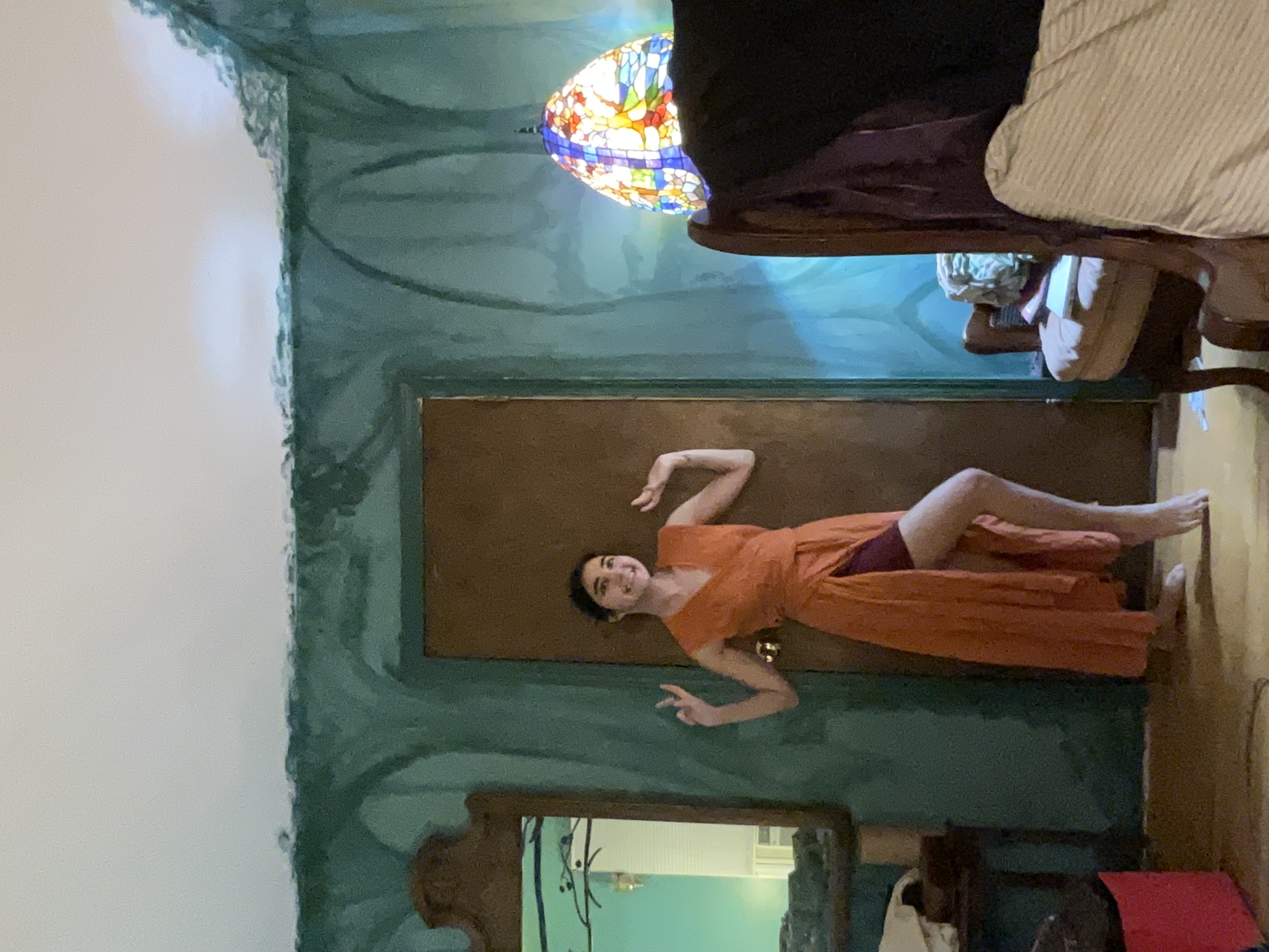 young woman in a long orange wrap dress poses in a room with teal walls. the room features a stainedglass lamp, an armchair, a mirror, and hardwood floors.  the walls appear to be in the process of being painted or repaired, showing some unevenness in the finish. the overall mood is casual and relaxed.