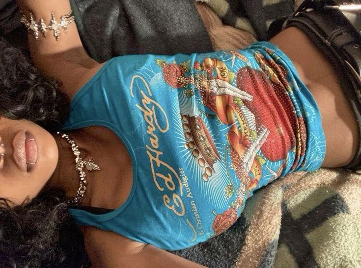 closeup image of a young woman lying down on a bed, wearing a turquoise ed hardy tank top with rhinestone detailing. the tank top features a graphic design. she is also wearing jewelry, including a necklace and bracelets. her dark, curly hair is visible, and she has several piercings, including a lip ring and septum piercing.  a portion of a blanket is visible underneath her.