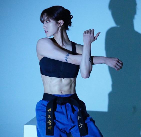 a woman with a muscular physique wearing a black sports bra and blue taekwondo pants with a black belt is captured in a studio setting. she is striking a pose, stretching her right arm across her body with her left hand gripping her right wrist. her hair is pulled back in a bun and she has a tattoo on her left bicep. the background is a light blue and there's a shadow of the woman cast on the wall behind her. the lighting is artificial, likely studio lighting.