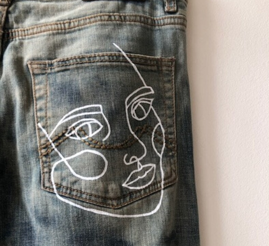 a closeup image of the back pocket of a pair of denim jeans. embroidered on the pocket is a minimalist line art portrait of a person's face. the embroidery is done in white thread, creating a striking contrast against the blue denim.