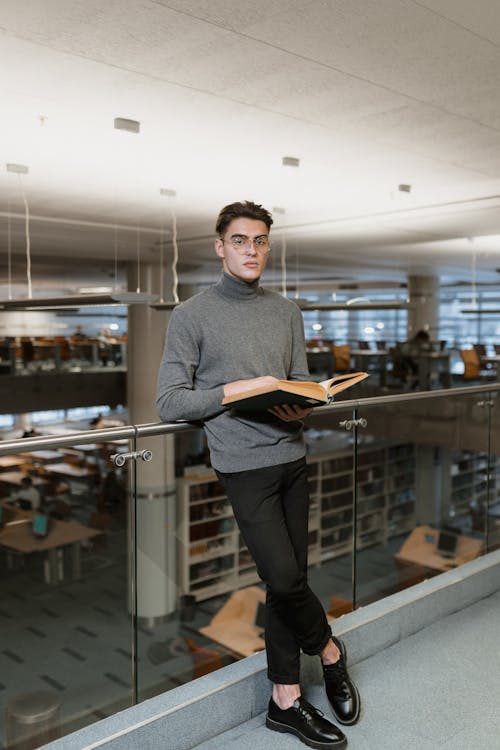 a man with glasses and a gray turtleneck sweater is standing in a library, leaning against a glass railing. he is holding an open book and appears to be reading. the library has shelves of books in the background and several tables and chairs.