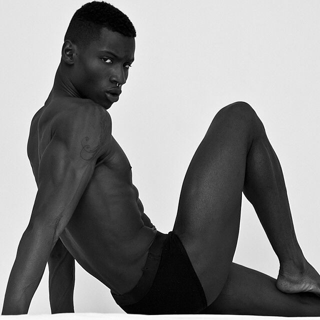 a black and white portrait of a young black man posing in a studio. he is sitting with his legs bent, his back facing the camera and his head turned to the side.  he is wearing black underwear, revealing his torso and muscular physique. his expression is serious and he is looking at the camera.