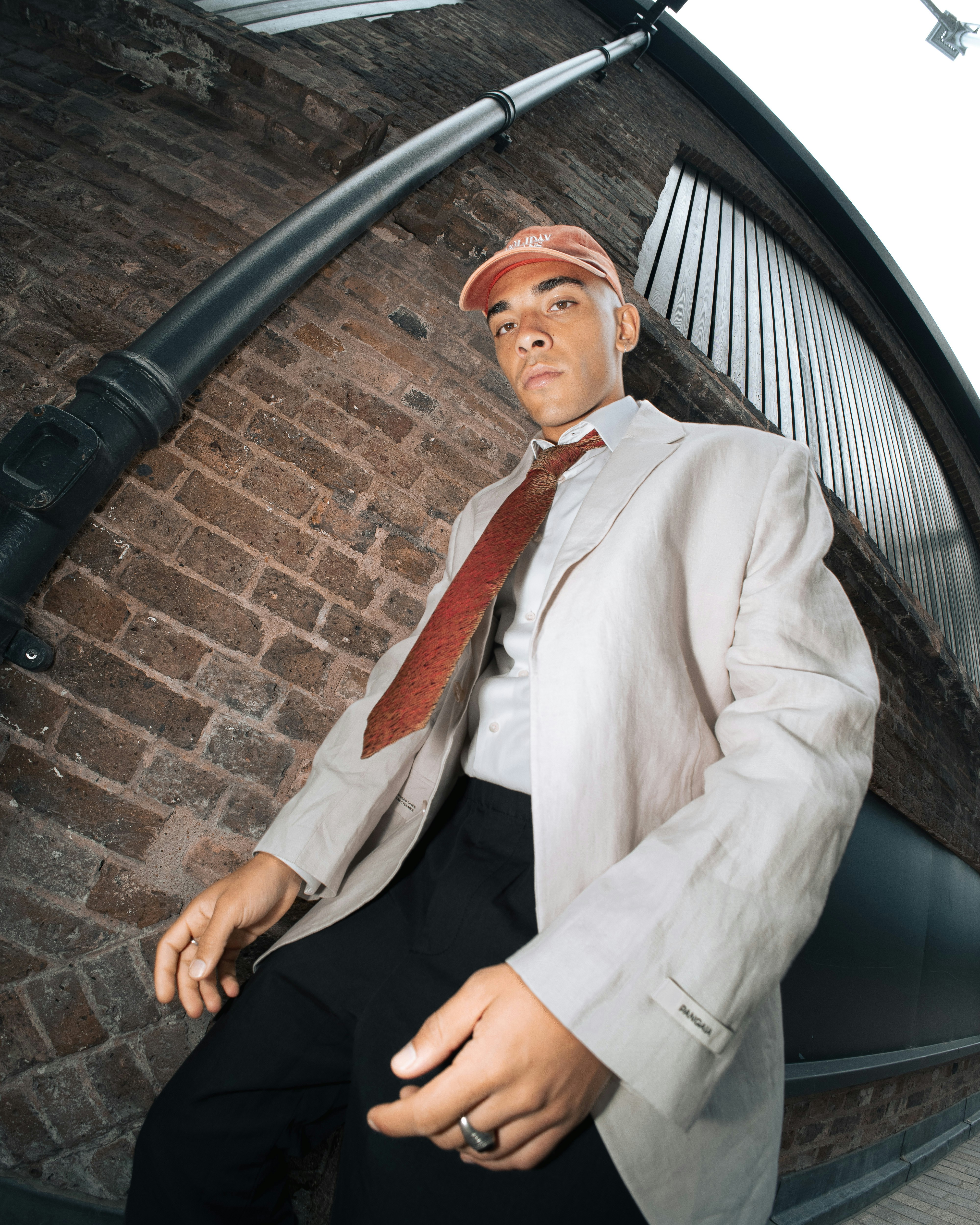 a young man in stylish clothing stands in a brickwalled urban alleyway. he wears a white blazer, red tie, black pants, and a red baseball cap. the image is taken from a low angle with a wideangle lens, creating a dynamic and slightly distorted perspective. the lighting is natural and overcast, casting soft shadows on the man and the brick wall behind him.