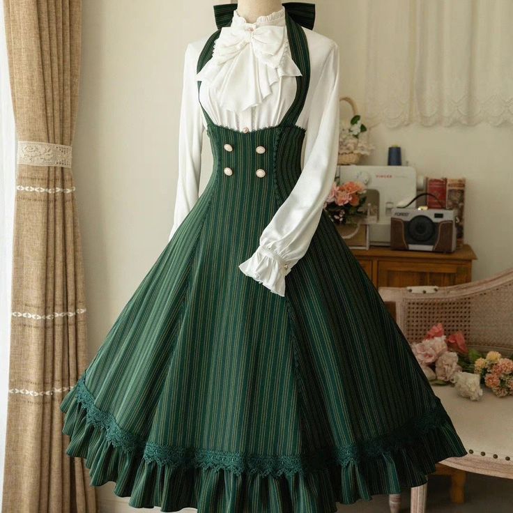 a fulllength photo of a woman wearing a green and white vintagestyle dress. the dress features a white blouse with a bow at the neck and a green, pinstriped jumper dress with buttons down the front. the skirt of the dress is full and has lace trim. the woman is posed in a room with floral curtains and furniture.