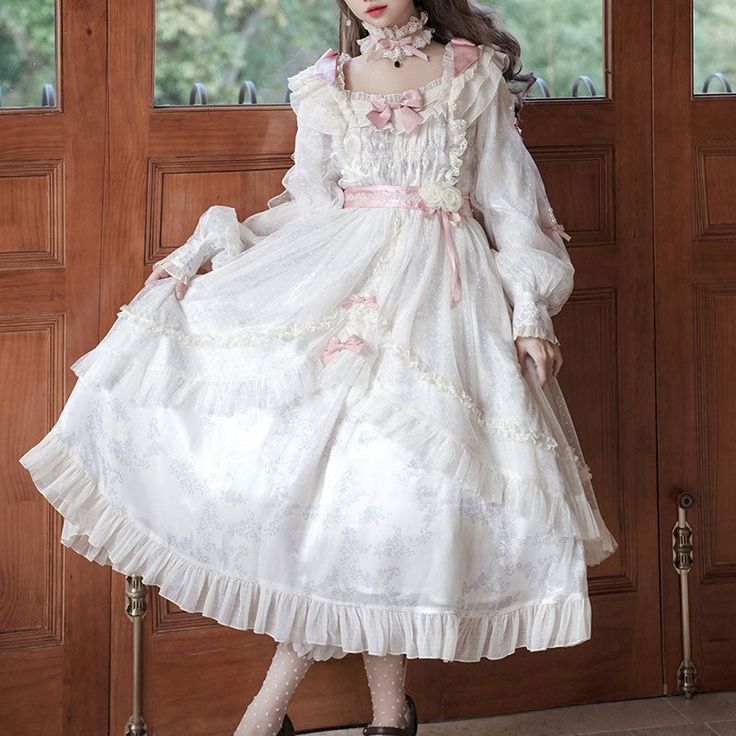 a girl is wearing a long white lolita fashion dress with ruffles and lace. the dress has pink ribbons and a pink flower. she is posing in an indoor environment.