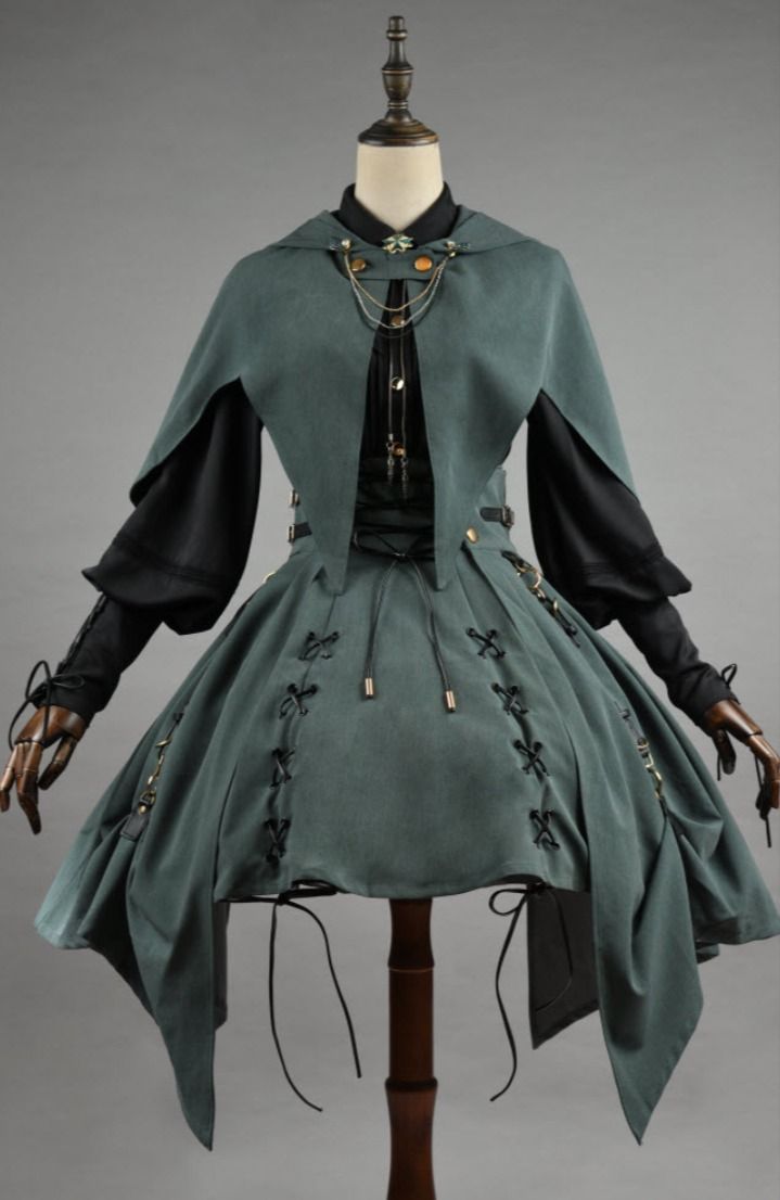 a green and black dress with a cape is displayed on a mannequin. the dress features black sleeves, a green skirt with laceup detailing, and an asymmetrical hem. the cape is attached to the dress and has a decorative brooch. the mannequin is positioned against a gray background.
