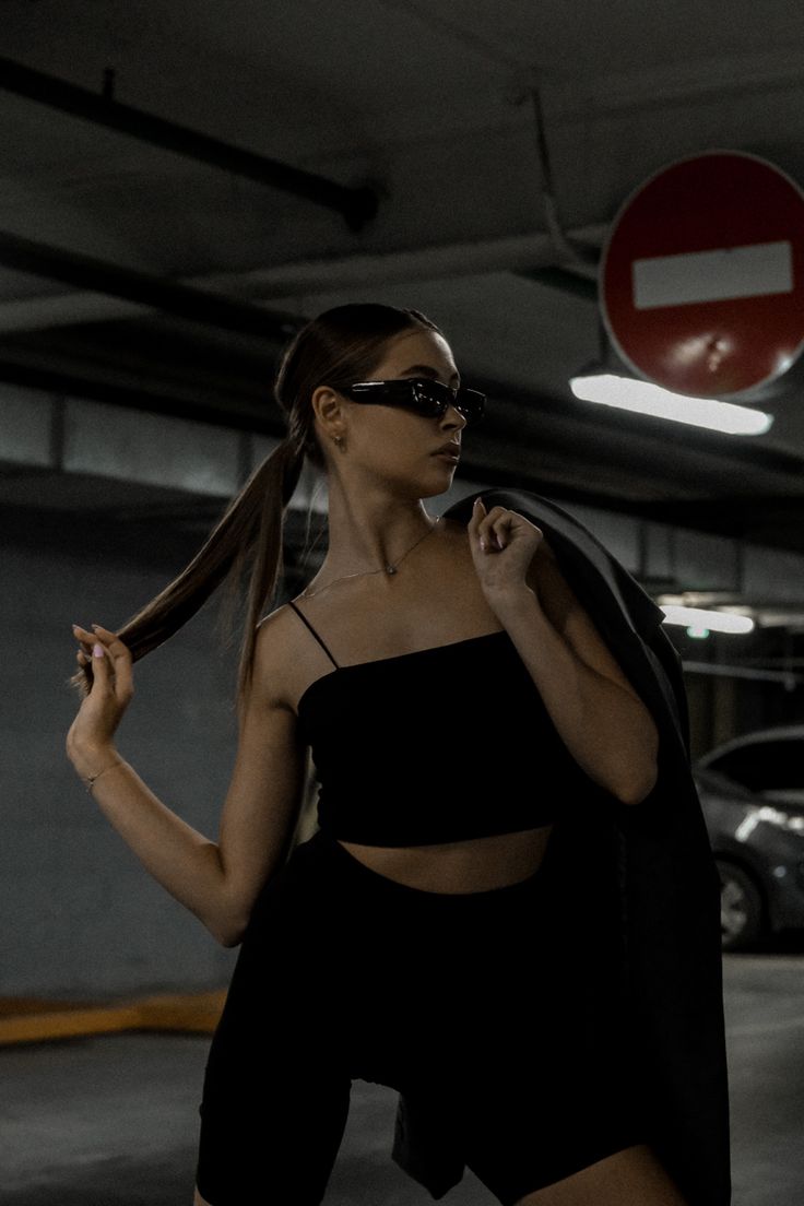 a woman in black sunglasses and a black outfit, poses with one hand on her hip and the other holding a black jacket.  she is in an urban setting with a car in the background. she looks confident and stylish.